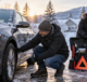 Driver checking a car tire and winter emergency supplies in a snowy driveway